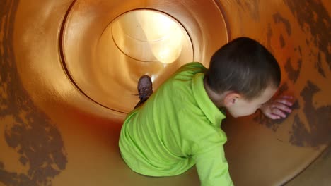 Little-boy-in-bright-green-shirt-slides-backwards-down-yellow-tube-slide