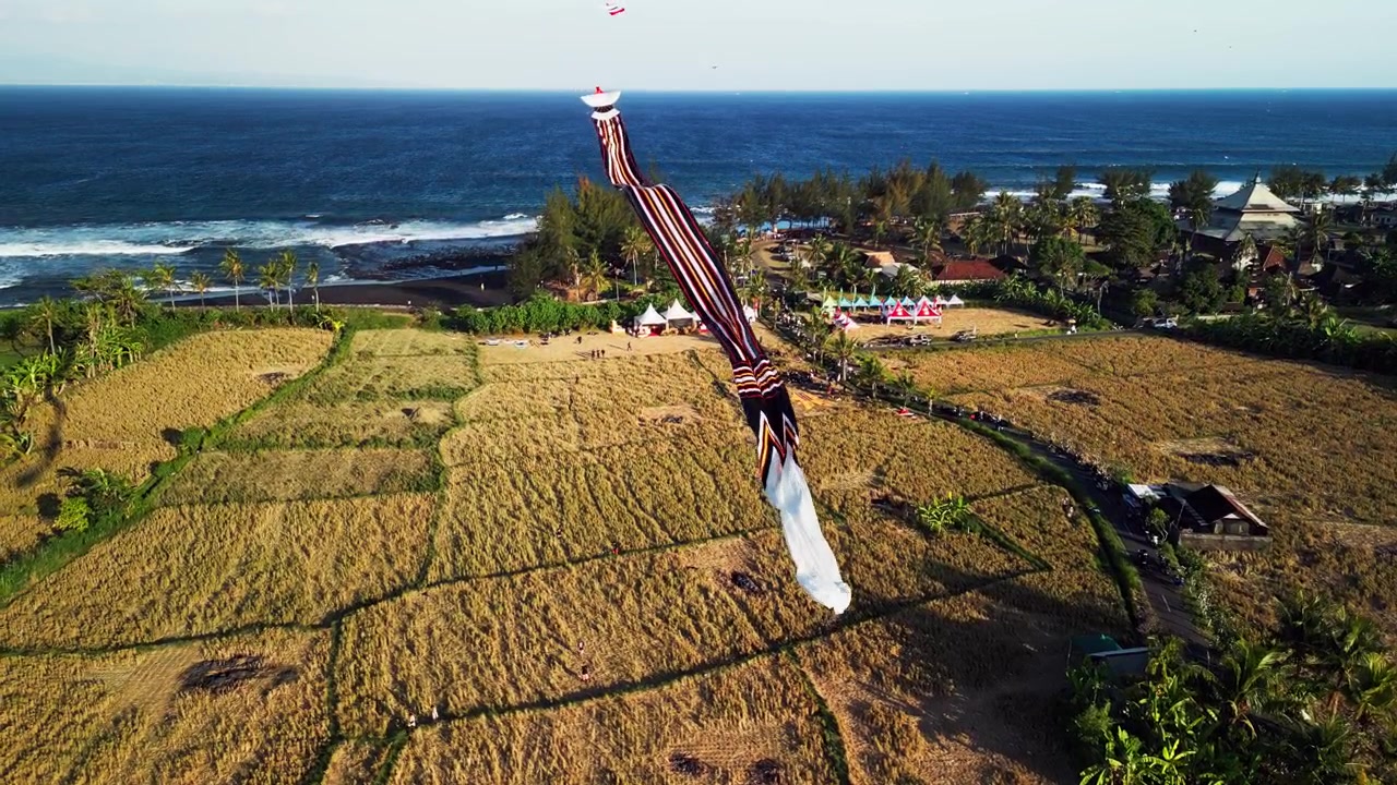 Aquilone gigante che si libra sopra un festival costiero accanto a risaie raccolte e campagna tropicale, mentre le onde dell'oceano si infrangono lungo la costa a Bali, Indonesia, riprese aeree con drone in ascesa.