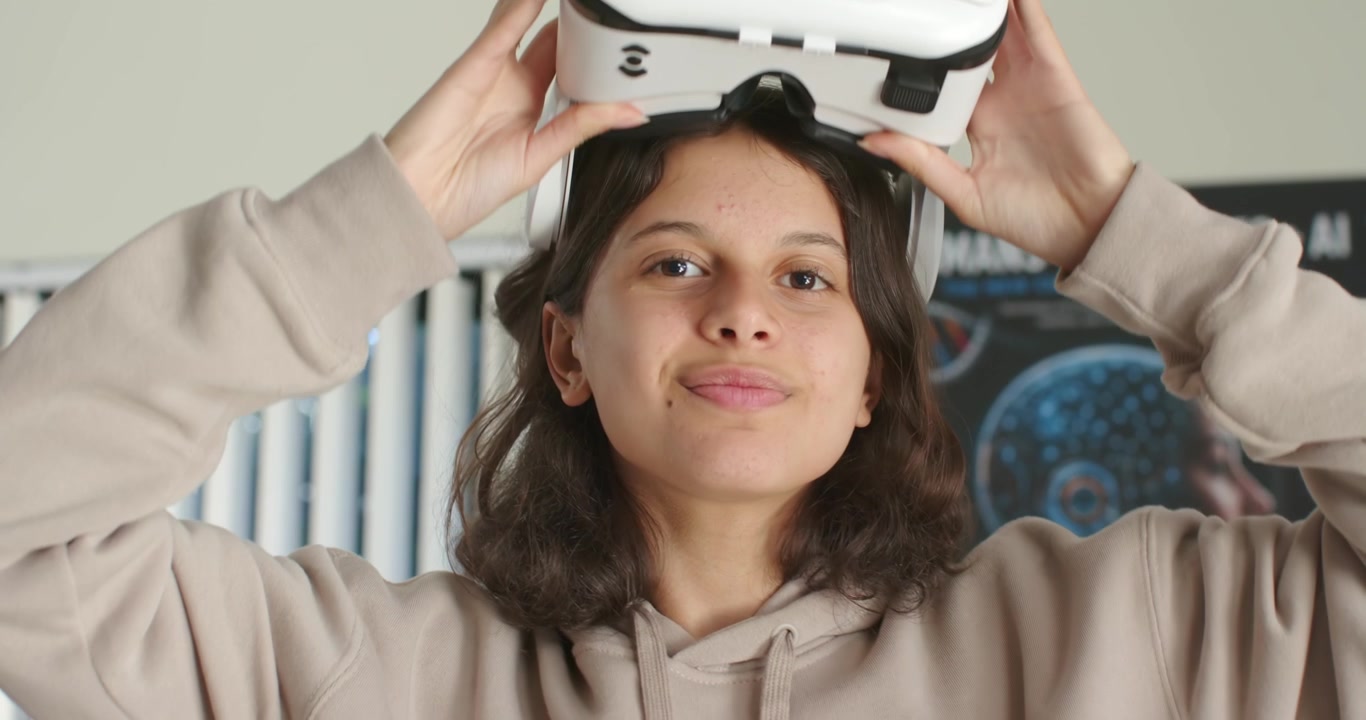 Ragazza adolescente afroamericana con felpa con cappuccio beige che afferra un visore VR bianco, testando la realtà virtuale in laboratorio.