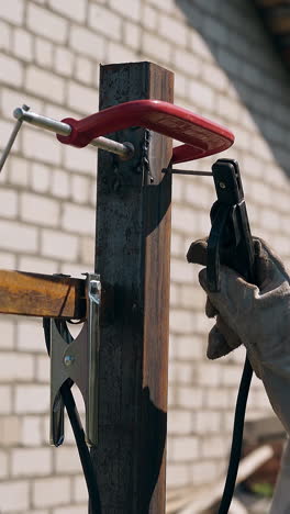 backside view man in protective mask welds small detail to rusty fence carcass top at brick building on day