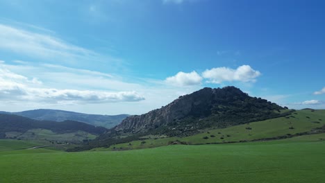 Flying-with-a-drone-over-a-green-meadow-with-some-spectacular-mountains-in-the-background-with-a-blue-sky-with-white-clouds