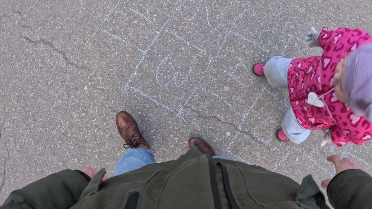 A parent’s perspective looking down at their child in a pink jacket, joyfully playing hopscotch on a sunny day.
