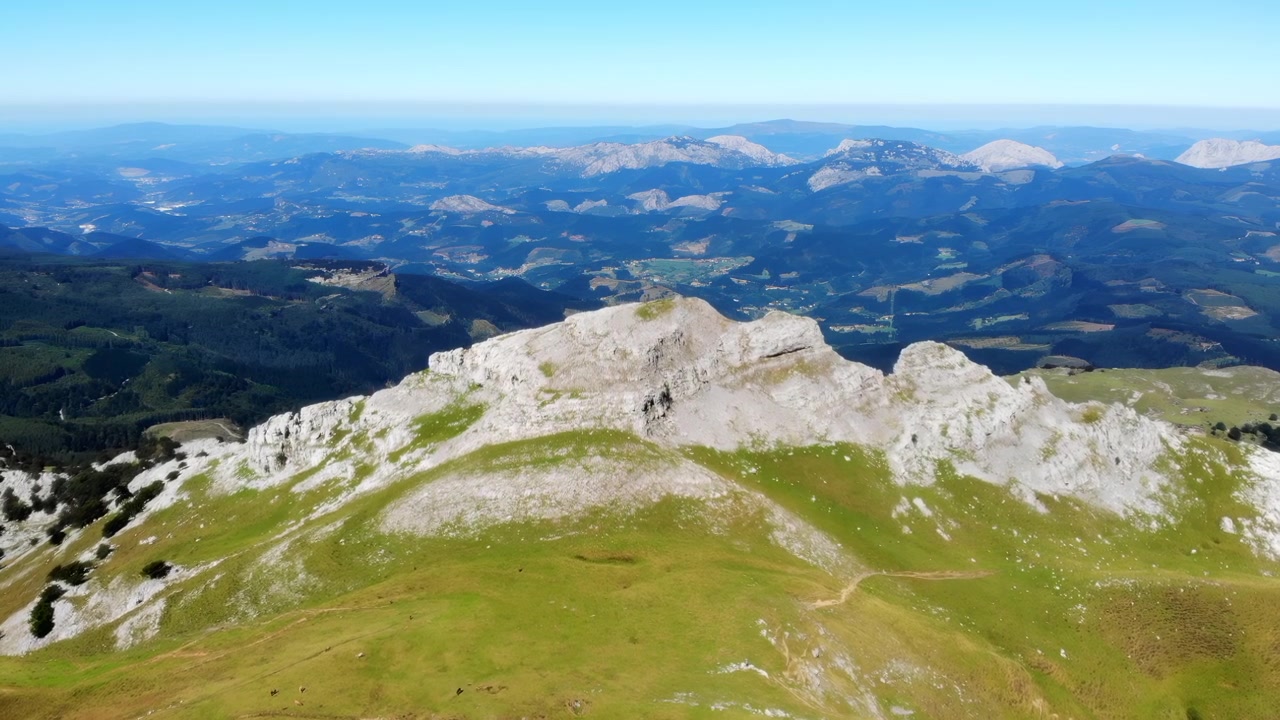 Ampia veduta aerea di una cresta montuosa rocciosa che si erge sopra dolci pendii alpini verdi, con cime stratificate distanti, valli aperte e un cielo blu chiaro e luminoso.
