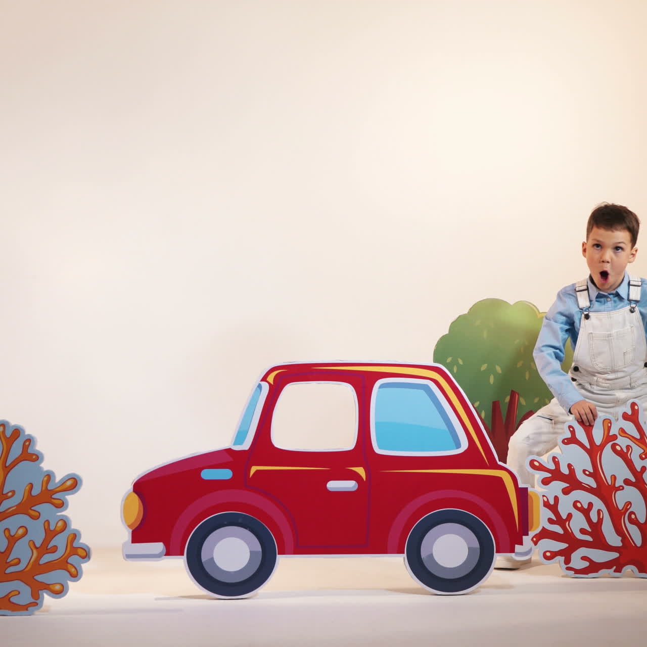 Cute little boy in white trousers comes to the children's room and shows surprised gestures. Childish studio with decor of toyful car, trees and bushes and kids playing in it.