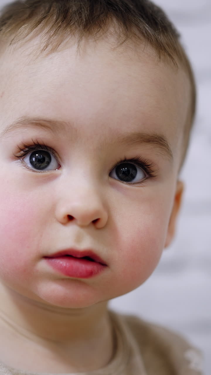 Adorable big-eyed baby boy with mousy hair. Portrait of a calm Caucasian kid focused on something. Close up. Blurred white backdrop. Vertical video