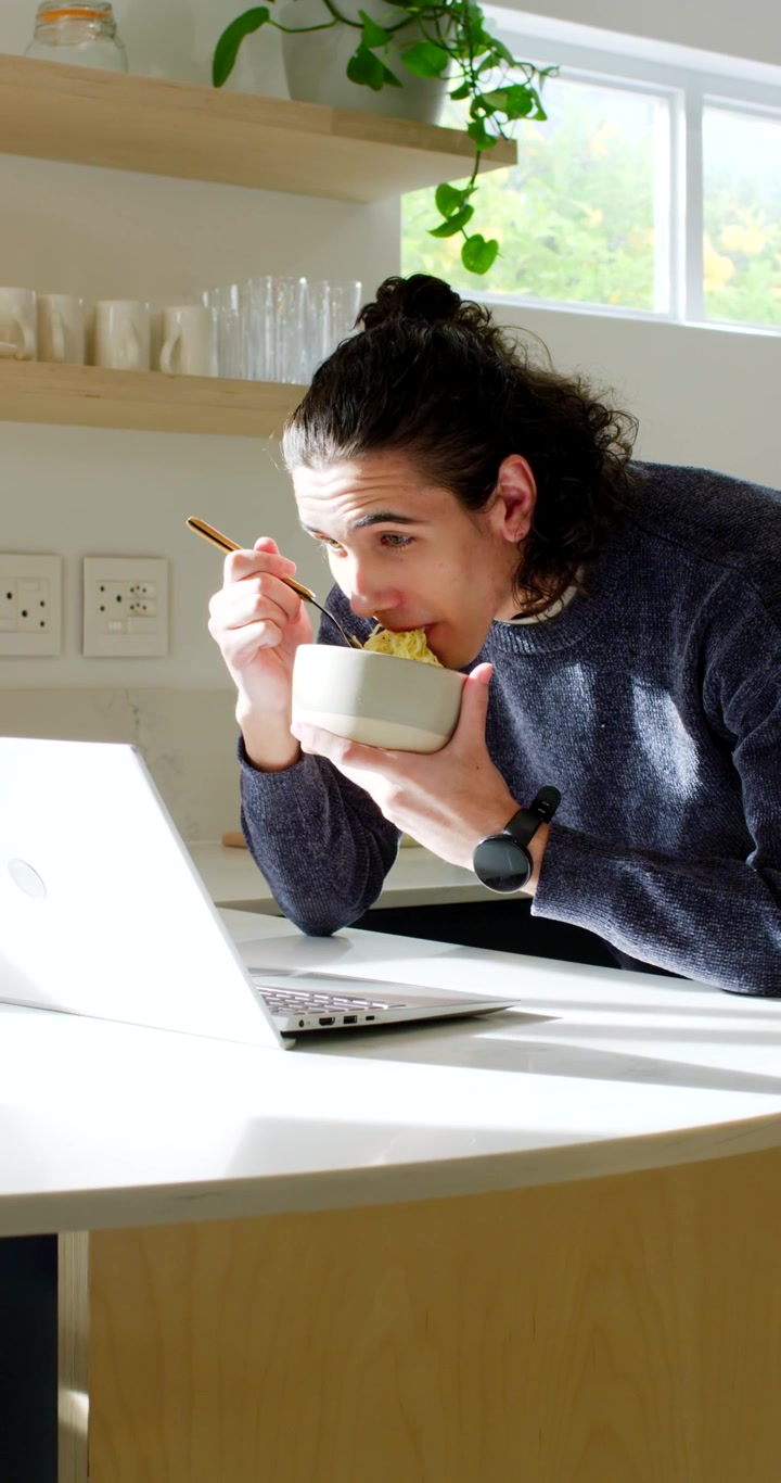 Vertical video: African American man eating noodles with bowl while working on laptop in kitchen