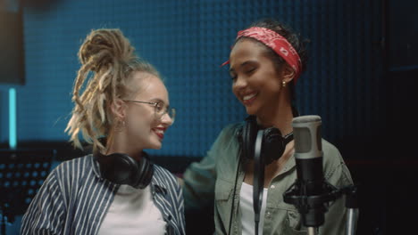 portrait of the two happy beautiful and stylish young women singers standing in the sound studio room at the mic, hugging and smiling cheerfully to the camera