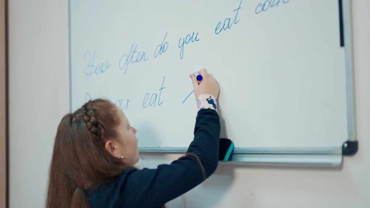 Schoolgirl writing on a blackboard. Side little girl in a school uniform writes with a marker on white magnetic board at primary school.