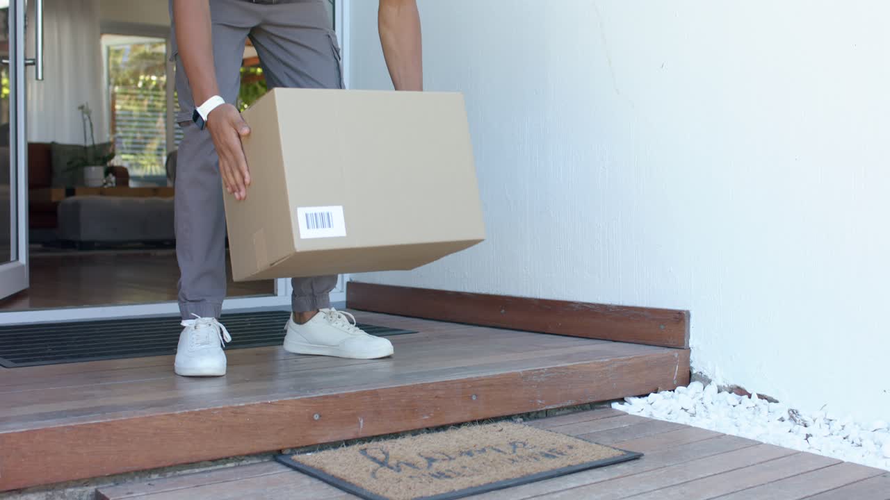 Man is stepping onto porch from ajar glass door, lifting cardboard box and carrying it inside