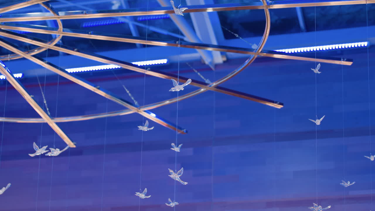 Interior decoration view showing suspended crystal birds under circular ceiling frame illuminated with blue light inside modern mall creating elegant artistic atmosphere with abstract pattern