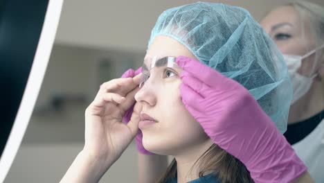 woman with brows ruler on face looks in mirror on tattooing