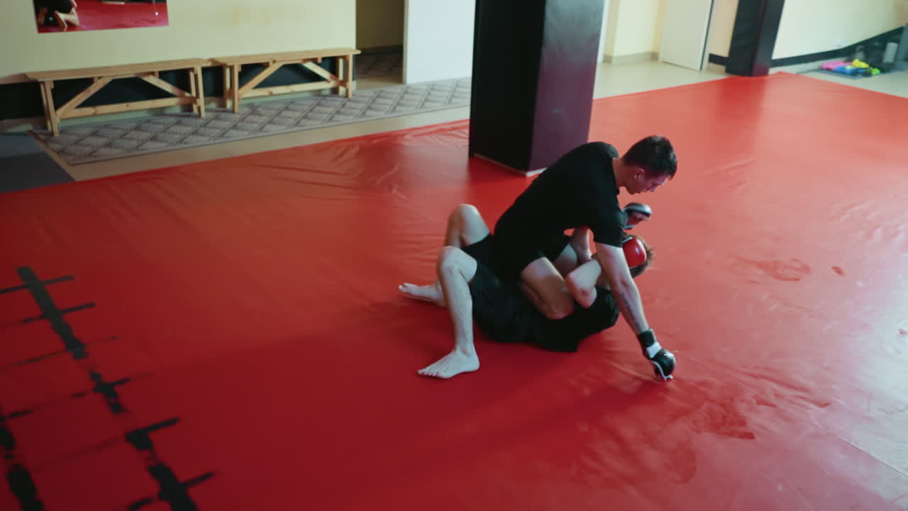 Battler executing ground control technique during combat training on red mats, demonstrating grappling, submission defense, endurance, strength,intensity in competitive practice