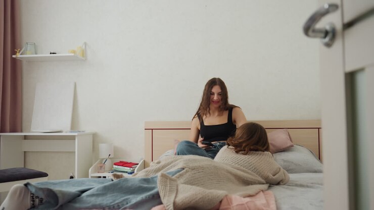 Madre caucasica entra silenziosamente nella camera da letto della figlia con un cardigan di lana, controllo delicato al letto mentre la figlia è seduta sul letto a leggere, luce mattutina soffusa, abbraccio confortante e calda atmosfera domestica