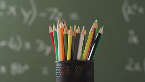 Video-of-cup-with-crayons-on-wooden-table-over-mathematical-formulas-on-blackboard