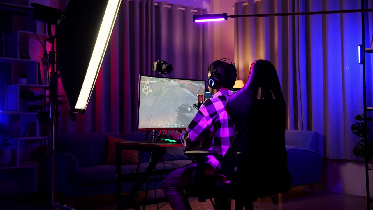 vista trasera de un chico asiático jugando a un juego en red en una computadora personal. videojuego de transmisión en vivo, escritorio iluminado por luz de tira led rgb
