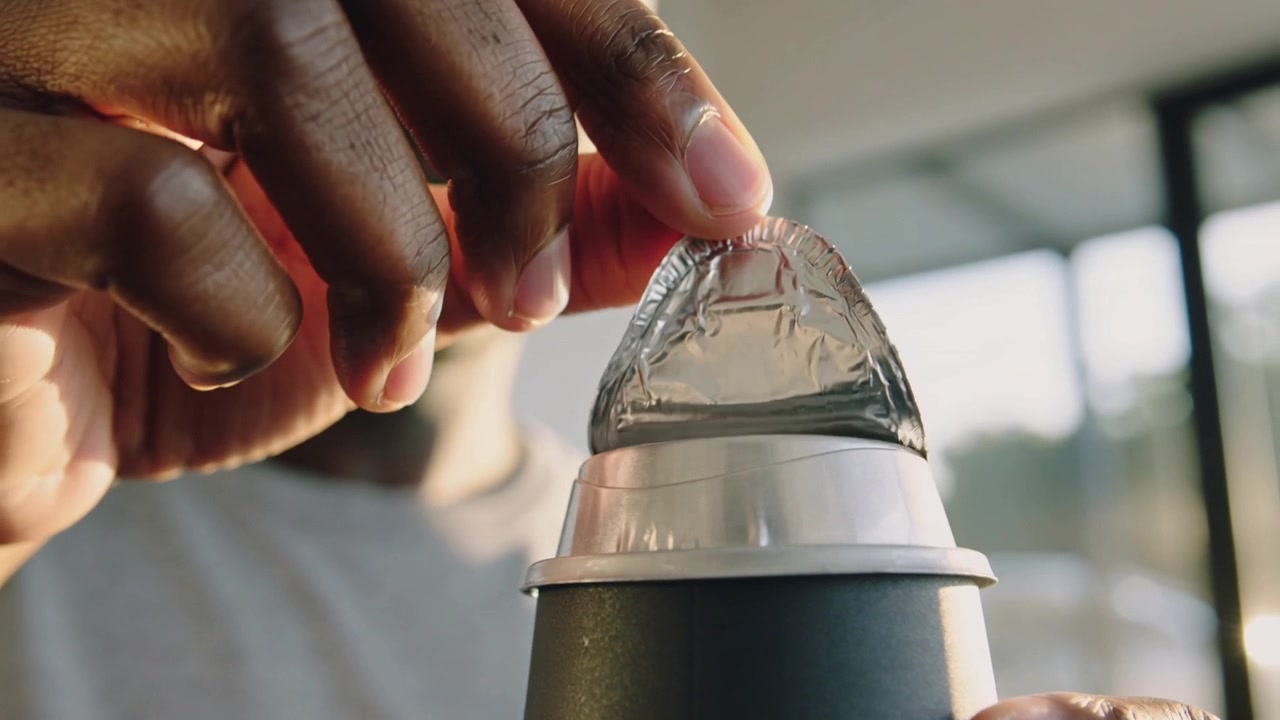 Peeling man hooking foil edge with finger, removing seal from cup by windows to access beverage