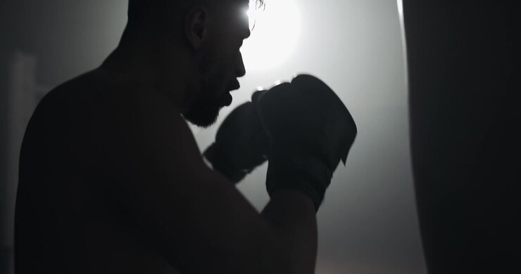 Silhouette di un pugile che si allena con un sacco da boxe in una palestra buia.