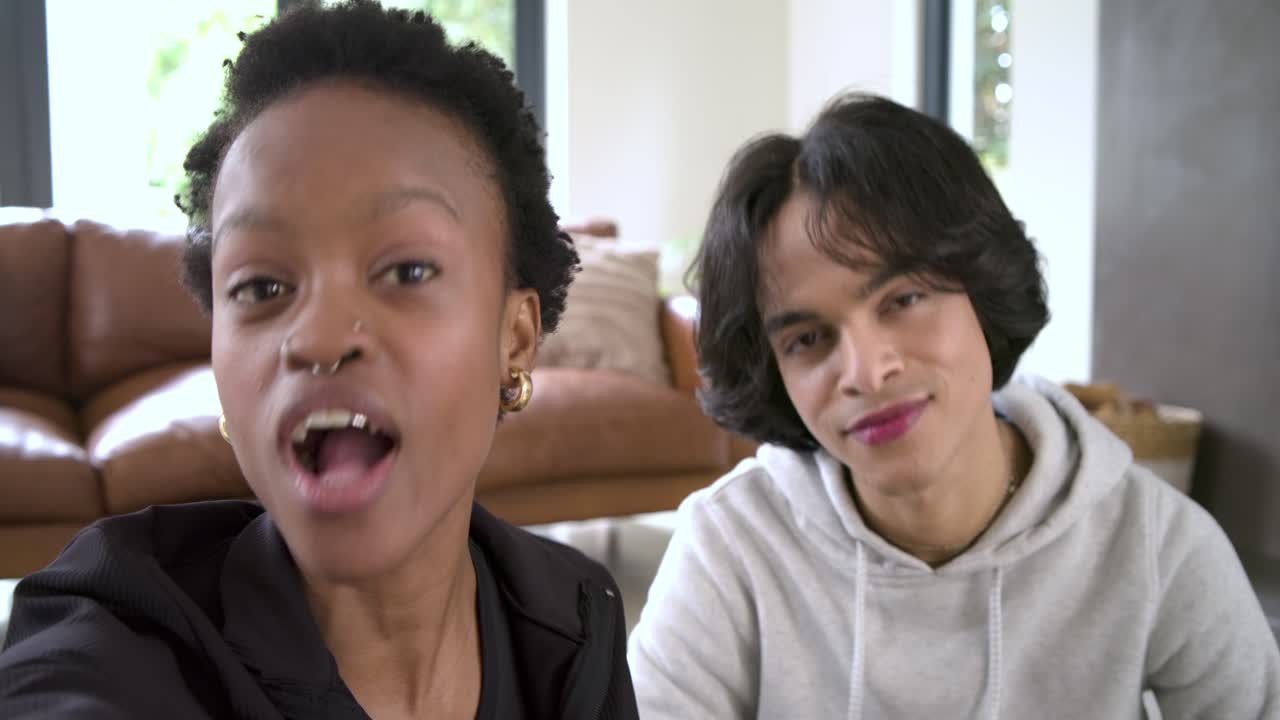 Un couple diversifié sur un canapé, face à la caméra, souriant, faisant signe de la main, puis parlant en riant avec une smartwatch