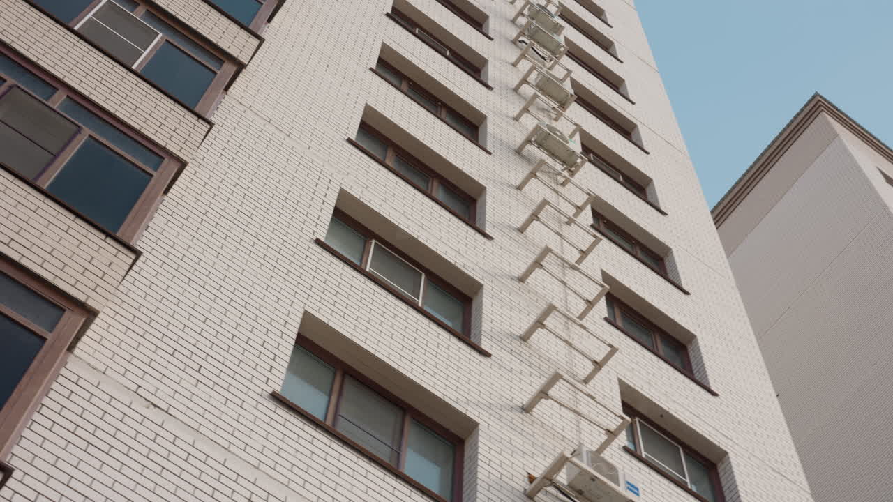 Worm eye view of tall brick apartment tower with stacked balconies and window air conditioners, perspective pulling upward through railing and surrounding urban architecture under soft sky light