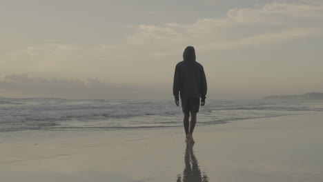 Ocean-waves-wash-into-young-barefoot-black-male-wearing-hoodie-walking-on-sandy-sunset-beach