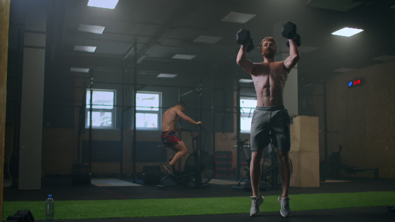 티셔츠를 입지 않은 운동 선수가 체육관에서 덤벨 운동을 합니다. 훈련 중에 무게를 들어 올립니다.