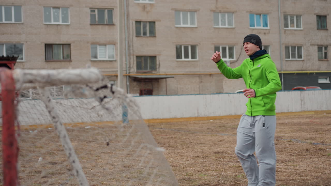Un jugador blanco camina de forma reflectante por un campo marrón después de entrenar; se ajusta el gorro con la mano; bloque de apartamentos al fondo; ambiente tranquilo, ritmo lento y aire fresco que transmiten fatiga e introspección.