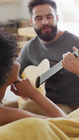 vertical video of happy diverse couple relaxing at home, man playing guitar to woman, copy space