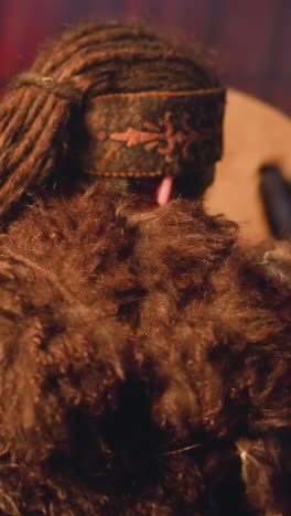 man with long dreadlocks and fur clothes plays shamanic tambourine at mystical ceremony in ail backside view. festival of altai native cultures