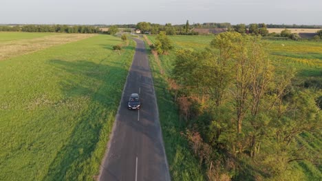 an aerial video of a car traveling along a straight rural road bordered by green fields and a calm lake. the scene captures the tranquility of the countryside under a clear blue sky