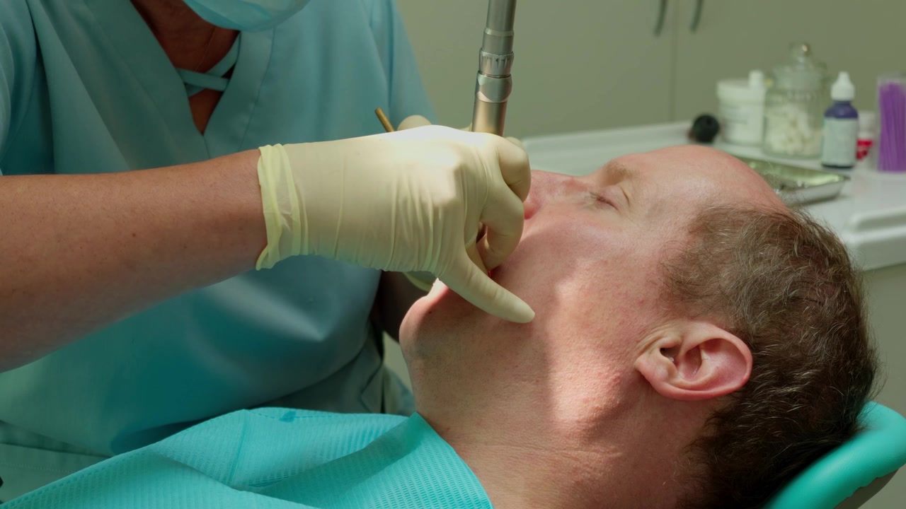 Una vista ravvicinata media mostra uno stomatologo professionista in camice che utilizza un preciso strumento di perforazione su un paziente maschio maturo che riposa sulla poltrona durante un controllo all'interno di uno studio dentistico.