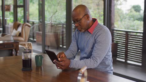 African-american-senior-man-sitting-at-counter-in-kitchen-concentrating,-using-tablet