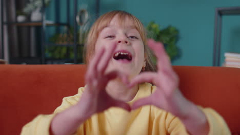 Smiling-child-girl-kid-on-home-sofa-looking-at-camera-makes-heart-gesture-demonstrates-love-sign