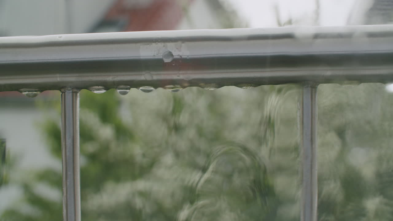 High-angle shot of raindrops falling and dripping from a French balcony railing, with a view into a typical German residential neighborhood in the background. Calm, overcast atmosphere