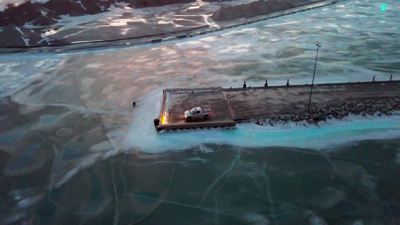 석양에 캐나다에서 겨울 동안 얼어붙은 호수로 둘러싸인 도크 끝에 앉아 있는 픽업 트럭 뒤에 궤도를 돌고 있습니다.