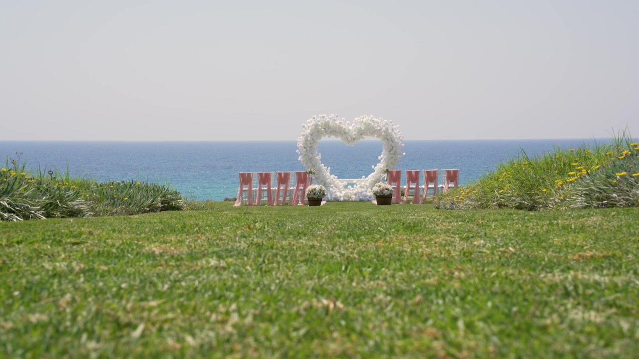 Folk forbereder sig til en bryllupsceremoni nær havet. Et hjerteformet blomsterarrangement står foran rækker af stole på grønt græs. Solen skinner, og vandet er roligt.