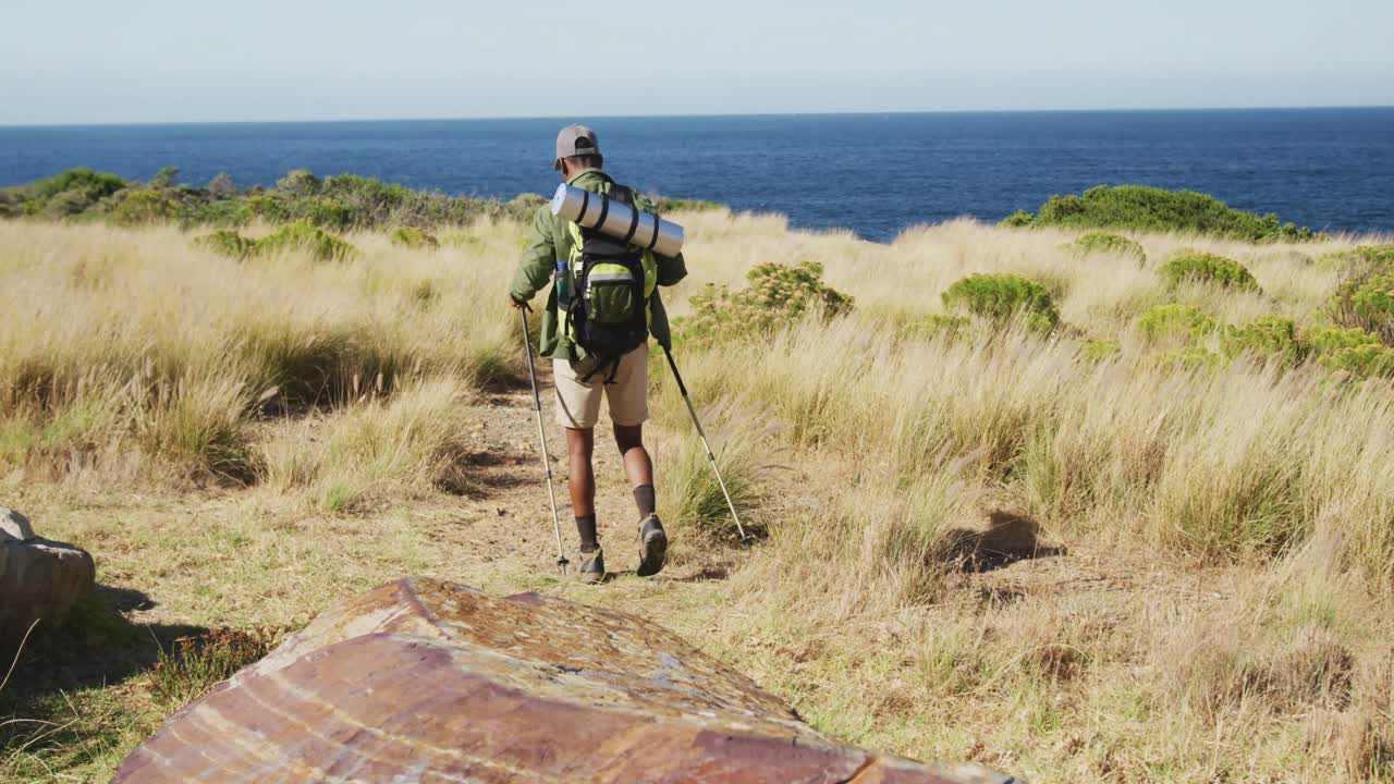 hombre afroamericano haciendo senderismo con palos de senderismo en el campo por la costa
