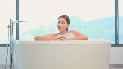 a pretty young woman in a bubble bath leans on the edge of her spa-like bathtub