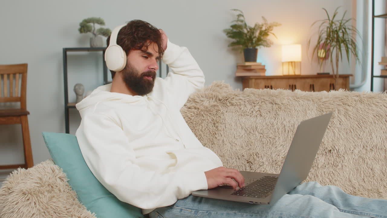 joven indio feliz y relajado con auriculares inalámbricos escuchando música usando una computadora portátil en el sofá