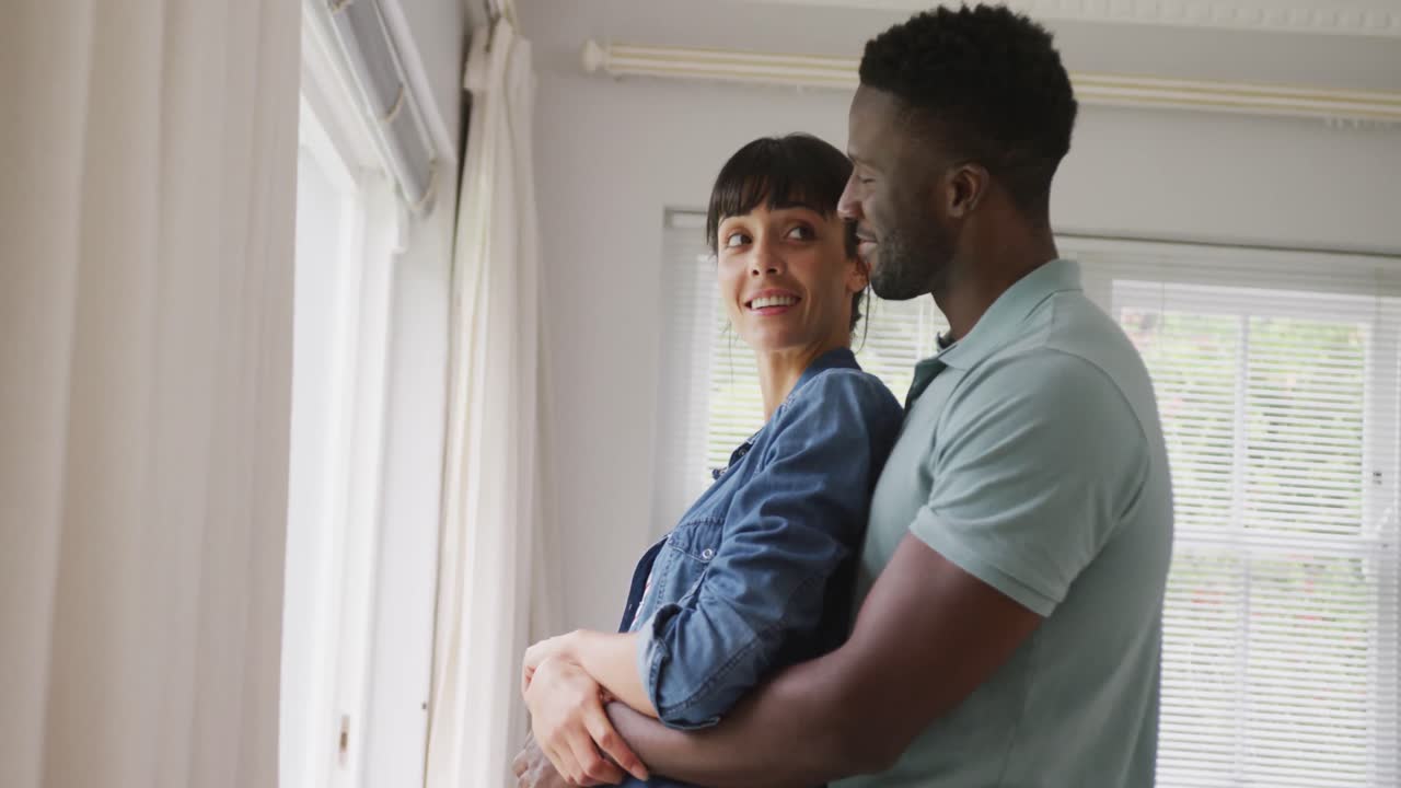 pareja feliz diversa usando chaqueta y camisa y abrazándose en la sala de estar