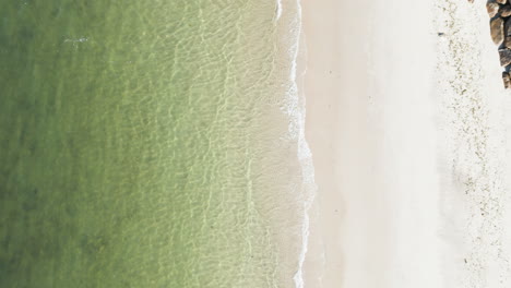 Drone-rises-above-calm-green-atlantic-ocean-waters-crashing-waves-on-sandy-shore