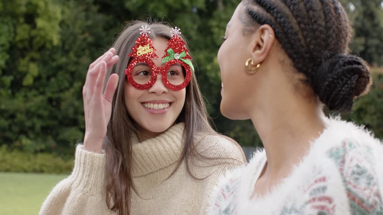 Diverse female friends lifting red holiday glasses laughing, adjusting for fun on garden bench