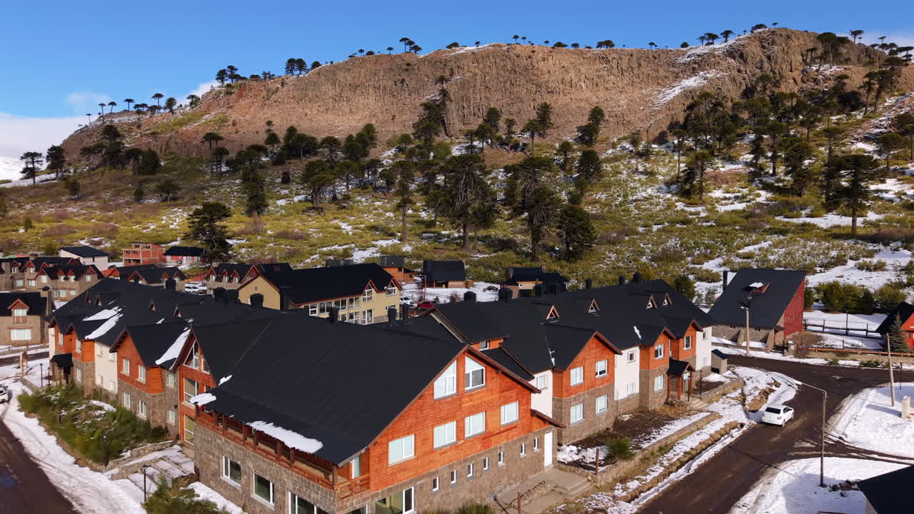 Caviahue, Neuquén, Argentina, showing colorful alpine-style houses surrounded by light snow, with Araucaria trees dotting the hillside under a clear blue winter sky, aerial dolly-in view