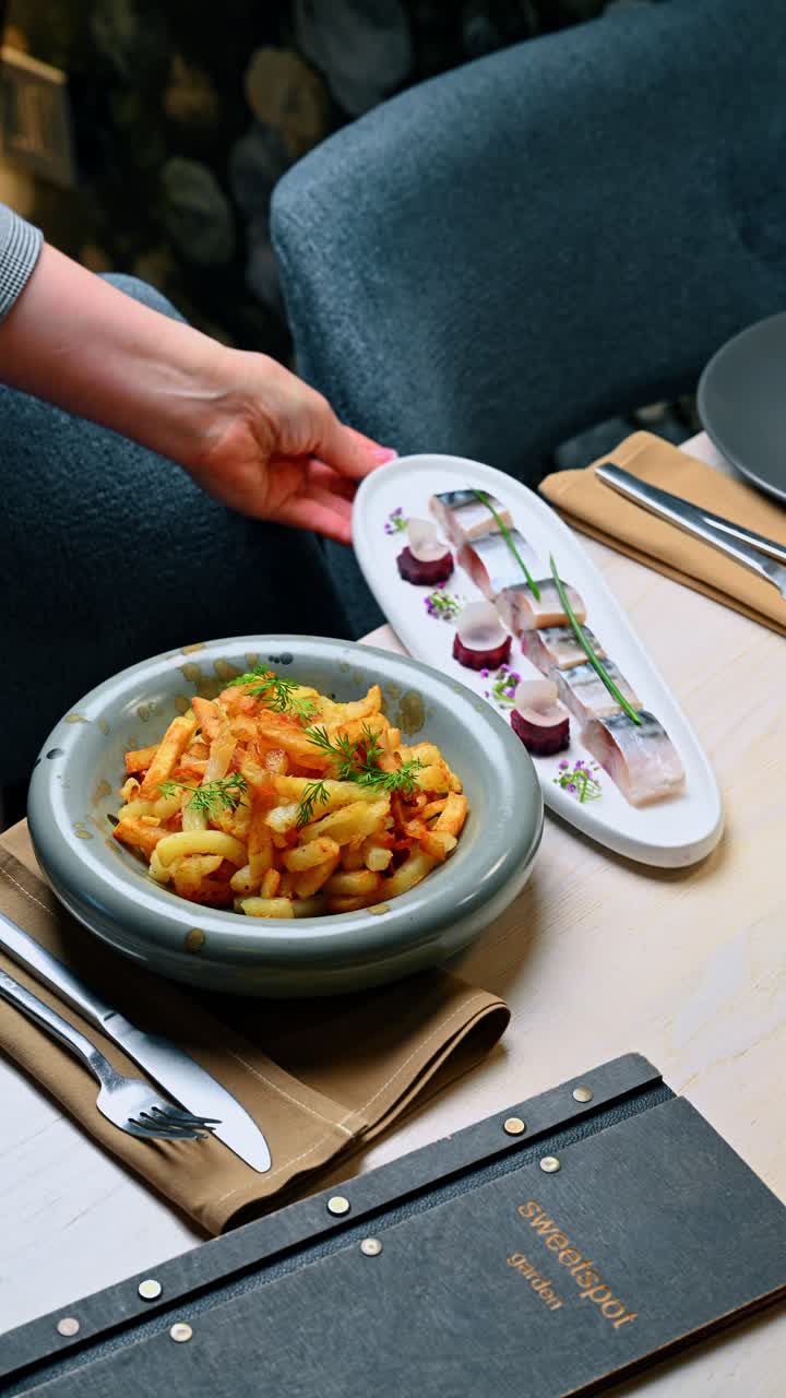 Tjener serverer en tallerken med sildemarinader og stegte kartofler. En hånd holder en lang hvid tallerken med fiskeanretninger ved siden af en skål med stegte kartofler på et bord.