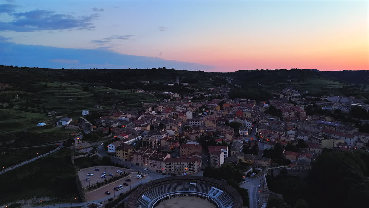 Reverse drone shot flying away from Brihuega, Spain, at sunrise. View reveals the full village, surrounding hills and historic bullring in warm early morning light. Peaceful rural atmosphere