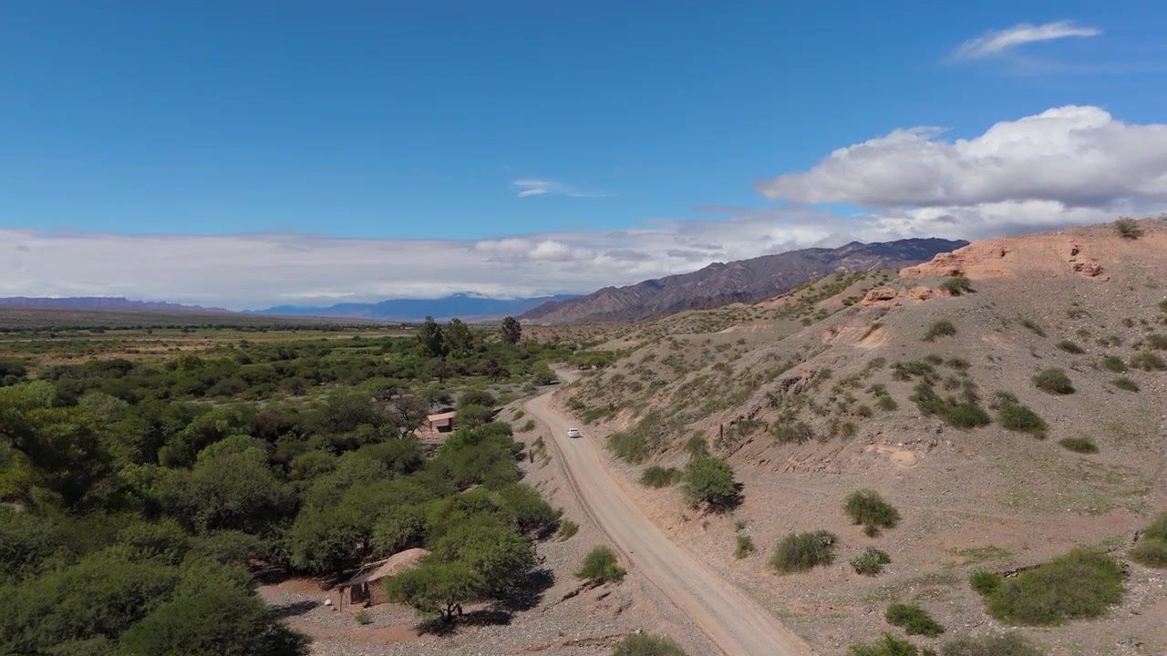 Dronebilde av en enkelt bil som kjører bort på en grusvei ved siden av en grønn dal nær Quebrada de las Flechas, Salta, luftfoto