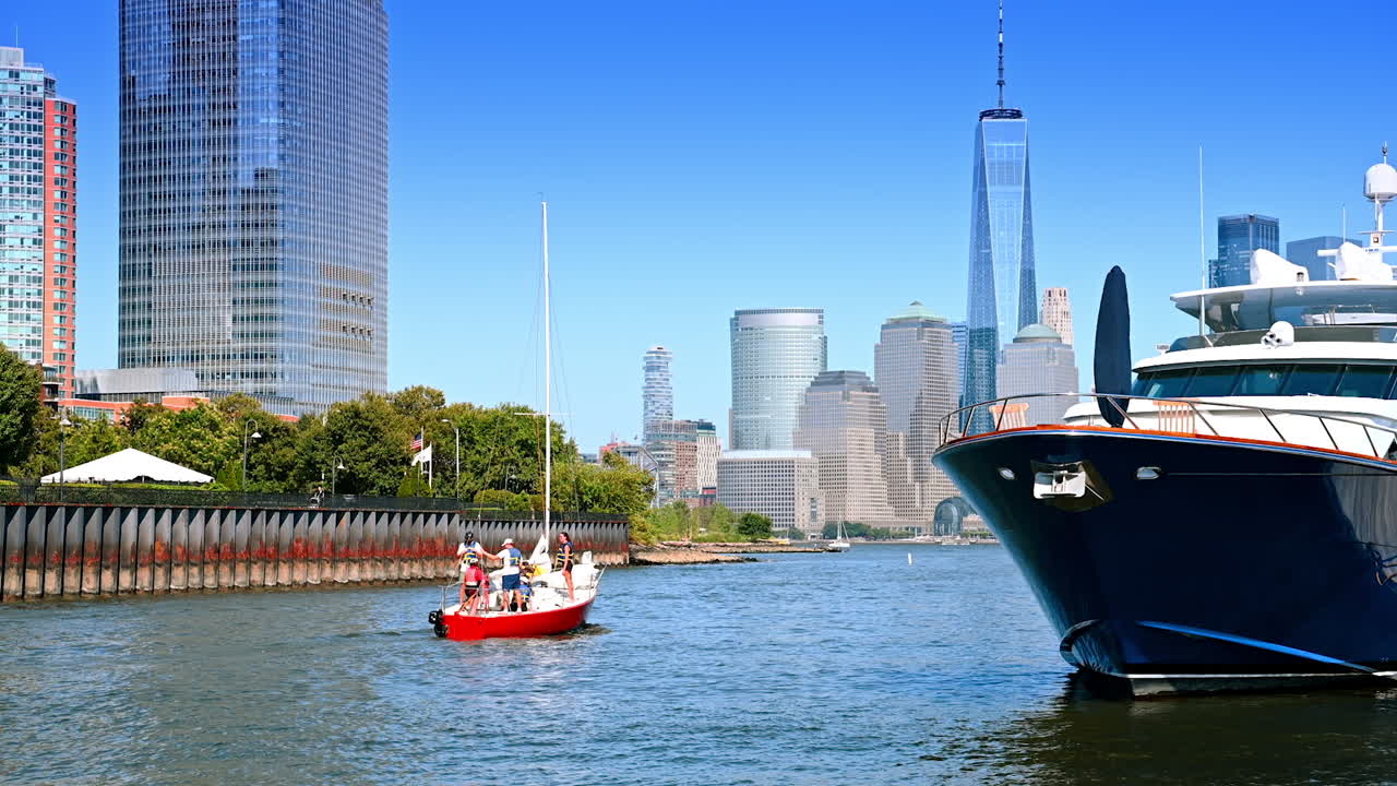 Jersey City, USA, 1 July 2025: Little red sailboat with a group of people moving by the river near big yacht. Sailing sport in Jersey City. Skyscrapers of Manhattan at backdrop