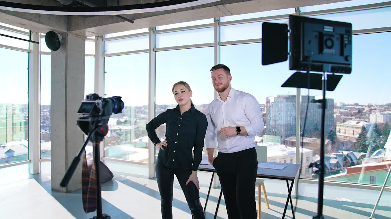 Young bloggers recording video about their business. Creative young couple shooting video in front of professional camera in office with panoramic windows.