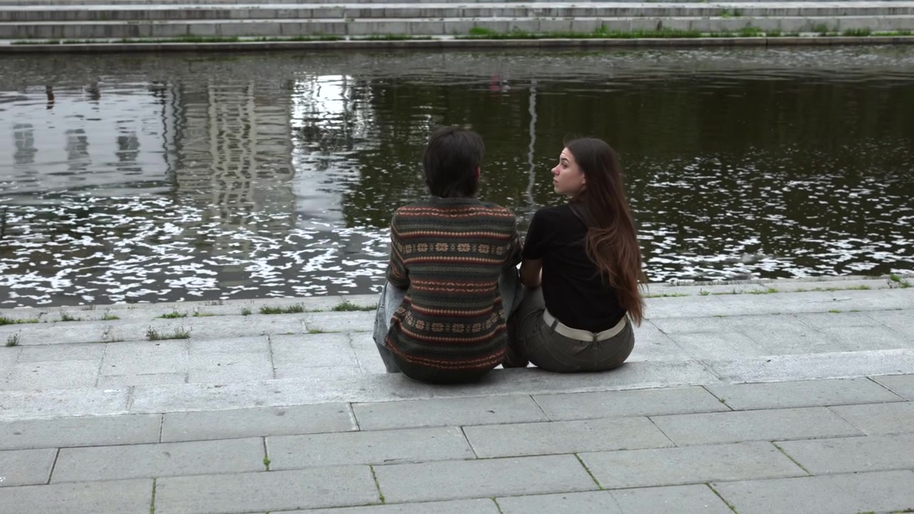 Two people sit side by side on tiered stone steps beside dark reflective canal, with one turning head toward companion while calm water, textured walls, grass, muted overcast light, and still urban