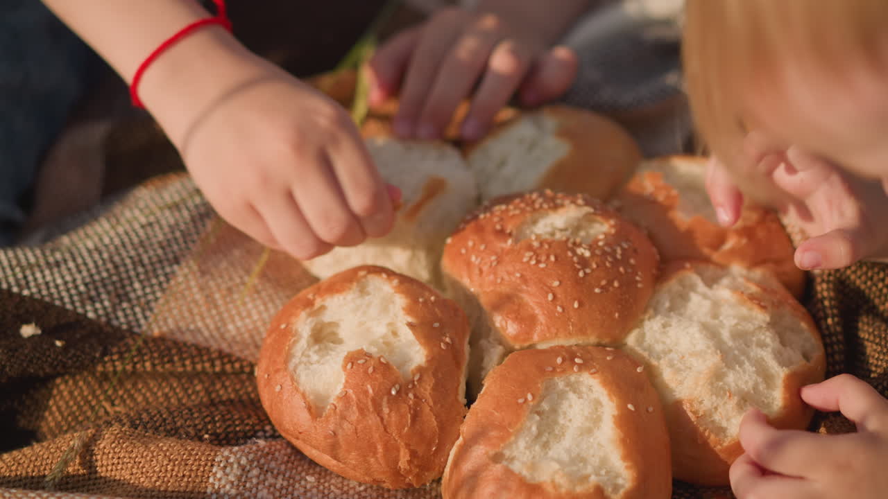 Close-up shot of children's hands pinching and eating freshly baked sesame bread rolls on a checkered picnic blanket. The scene captures a simple and peaceful outdoor moment