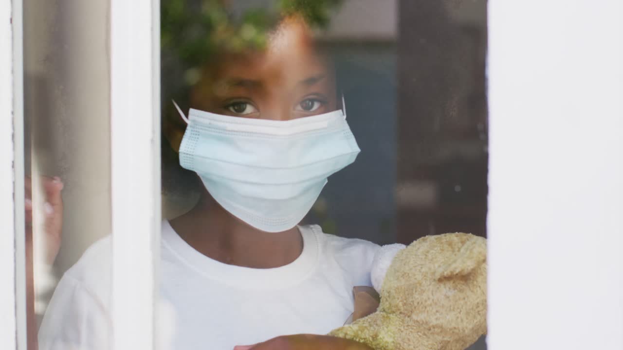 아프리카계 미국인 소녀가 얼굴 마스크를 쓰고 테디 베어를 들고 창문을 통해 바라보고 있다